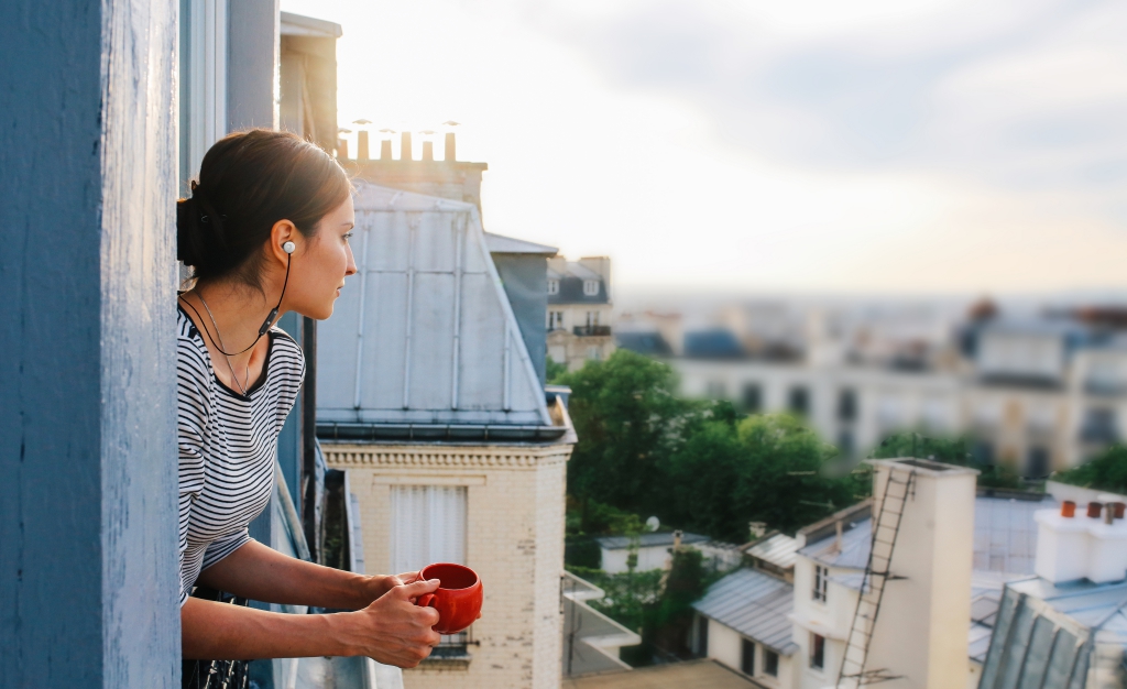 Vintage toned image of a young woman relaxing, drinking coffee, on the small balcony - window of her beautiful apartment on Montmartre, Paris. Taken in the magic hour just as the sun sets down over Parisian city scape in the background and the sunrays paint her hair bright.